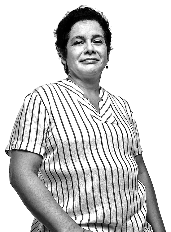 Una imagen en blanco y negro de una mujer de pie, con una expresión serena y segura. Luce una blusa de rayas verticales y tiene el cabello corto y rizado. Su postura sugiere confianza, mirando ligeramente hacia un lado. El fondo es completamente negro, lo que resalta su figura y la prenda que lleva puesta.